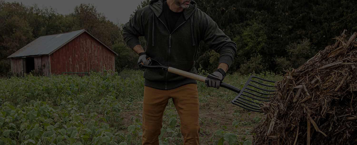 A field worker spreads forage in the middle of a lush field with a pitchfork. A red barn appears in the background next to a woods.  