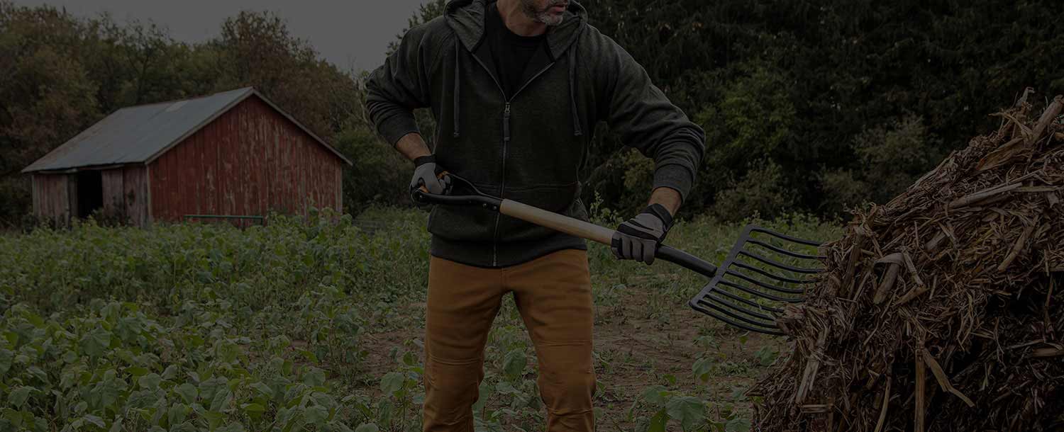 A field worker spreads forage in the middle of a lush field with a pitchfork. A red barn appears in the background next to a woods.