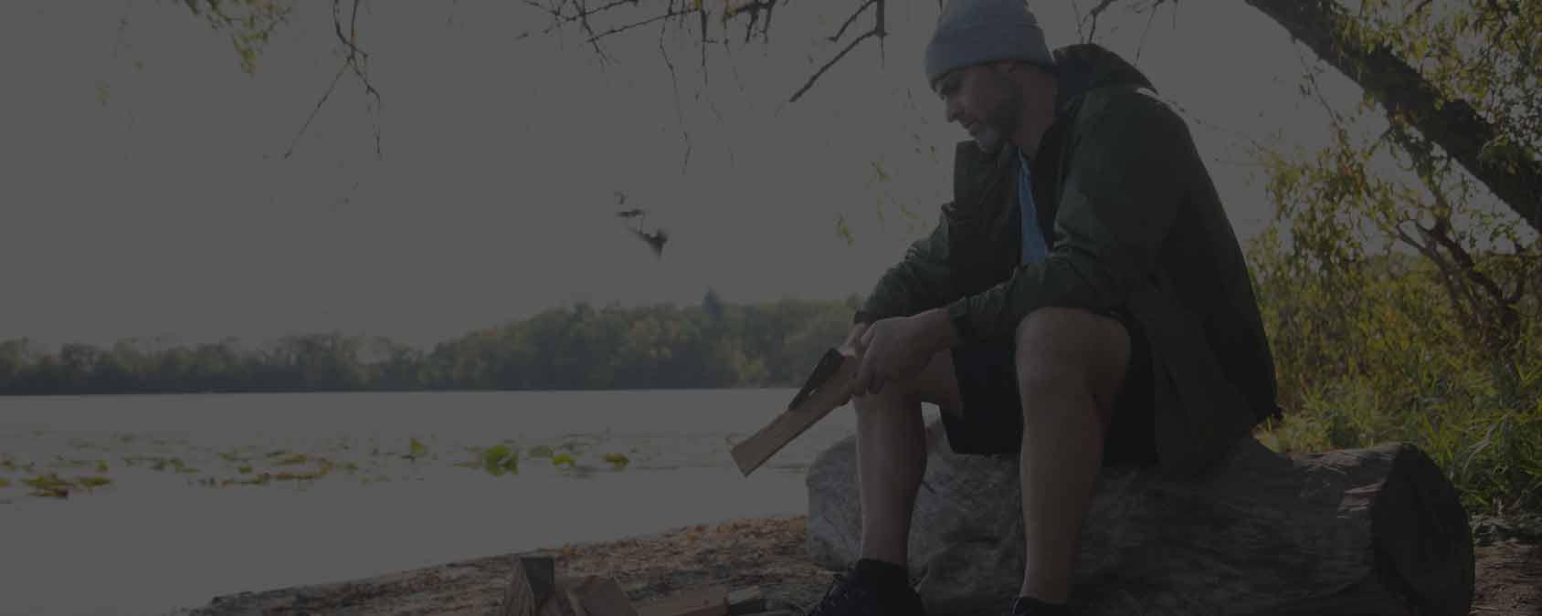 A man sits outdoors by a campfire and flowing river