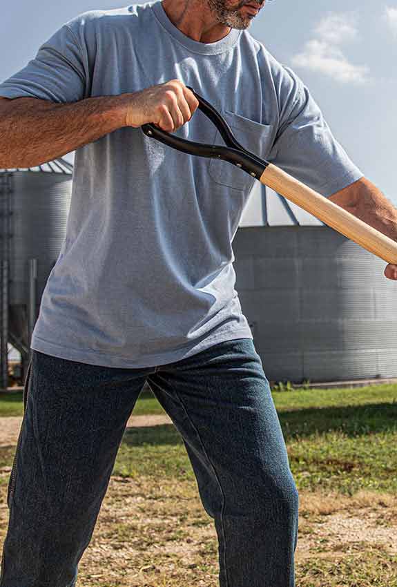 A man in a light gray Work n’ Sport Heavyweight Pocket Tee shovels corn. There is a grain storage silo behind him.