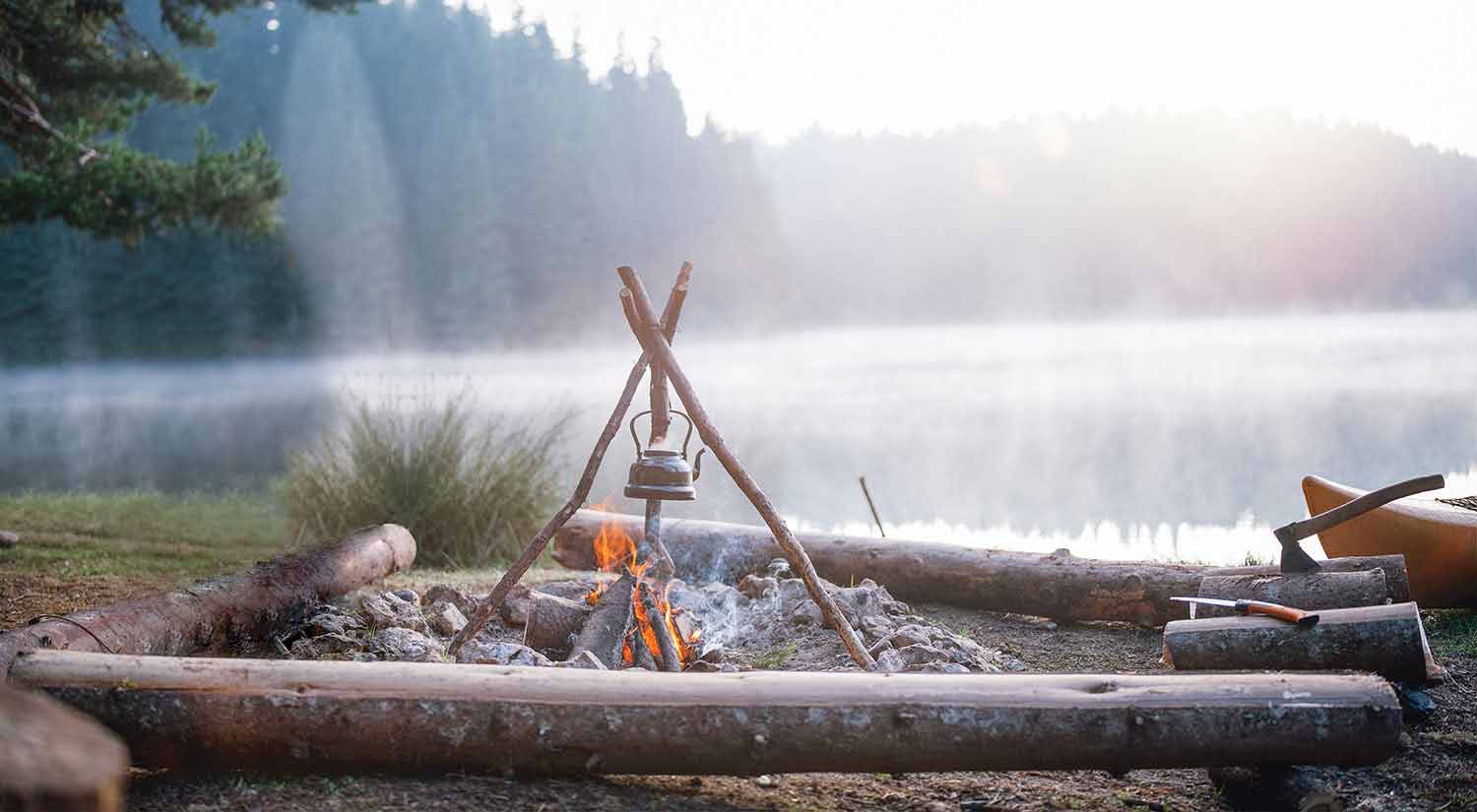 A kettle hangs over a roaring campfire with a beautiful, misty lake in the background. 
