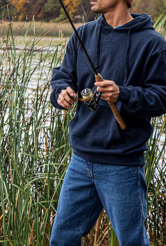A fisherman wearing a Work n’ Sport Men’s Hooded Sweatshirt stands among the reeds, poised to cast his line.