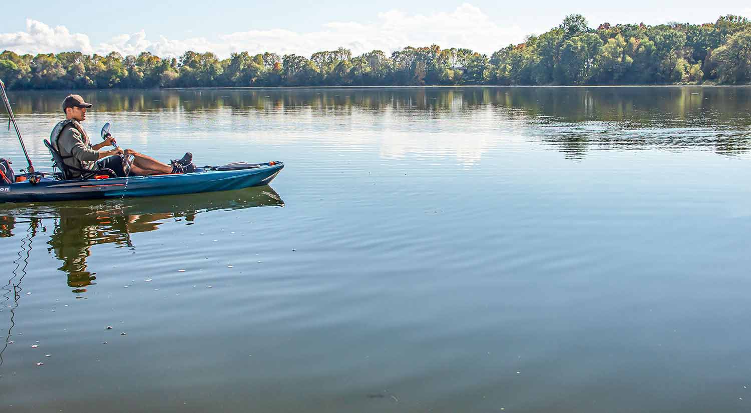 A kayaker paddles slowly through a placid lake with a blue sky above and a verdant treeline in the background 